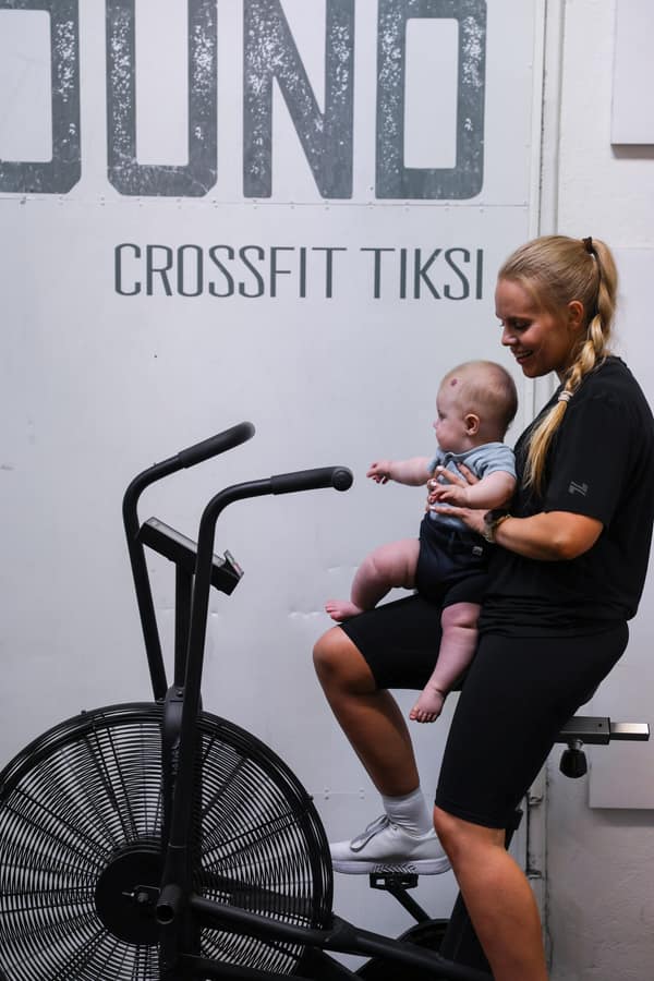 Valmentaja istuu ilmapyörällä vauva sylissään, seinässä teksti CrossFit Tiksi.