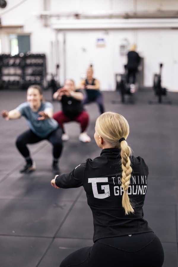 Valmentaja Training Ground -takissa ohjaa kyykkyjä tekevää ryhmää.