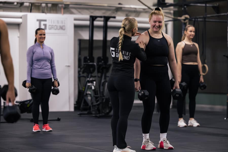 Valmentaja Training Ground -takissa juttelee hymyilevän treenaajan kanssa käsipainotunnilla, taustalla muita hymyileviä treenaajia.