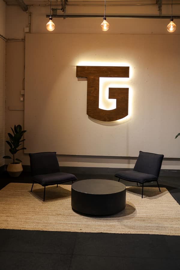 Salin aula: valaistu puinen TG-logo seinällä, kaksi nojatuolia, pyöreä pöytä ja viherkasvi.